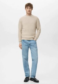 Lichtbeige geribbelde trui, relaxte blauwe spijkerbroek en zwarte loafers. Model staat recht vooruit gericht tegen een effen achtergrond.