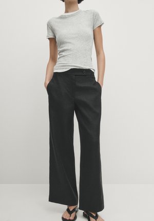 Personne portant une chemise à manches courtes gris clair, un pantalon large noir avec poches et des sandales noires à bouts ouverts sur un fond uni.