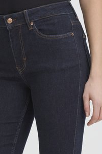 Detailní záběr tmavě modrých džín s měděnými knoflíky, žlutým šitím a rukou opřenou vedle stehna.