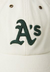 Gorra de pana blanca con un logotipo bordado en verde de los "A's". La tela tiene una textura estriada y cuenta con una arandela de metal en la parte superior.