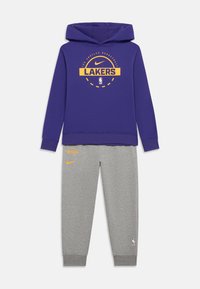 Lilla hættetrøje med "LAKERS" logo i gul, parret med grå sweatpants med orange accenter og NBA logo patch på benet.