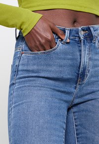 Jeans de mezclilla azul claro con diseño de cinco bolsillos, costuras visibles y cierre de botón metálico. Combinados con una camiseta verde de manga larga ajustada.