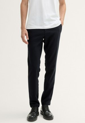 Man die zwarte slim-fit broek, zwarte leren veterschoenen en een wit T-shirt draagt, staand met één hand in de zak.