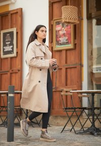 Manteau beige à double boutonnage, leggings noirs et baskets montantes marron avec des lacets blancs, le tout devant une porte en bois.