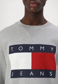 Sivý mikina s rebrovaným výstrihom, s odvážnym dizajnom loga v námorníckej modrej, bielej a červenej farbe, s textom "TOMMY JEANS".