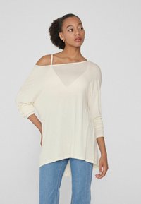 Hellcremefarbenes Langarmoberteil mit off-shoulder Design, locker sitzend und mit weitem Ausschnitt, kombiniert mit hoch taillierten blauen Jeans.