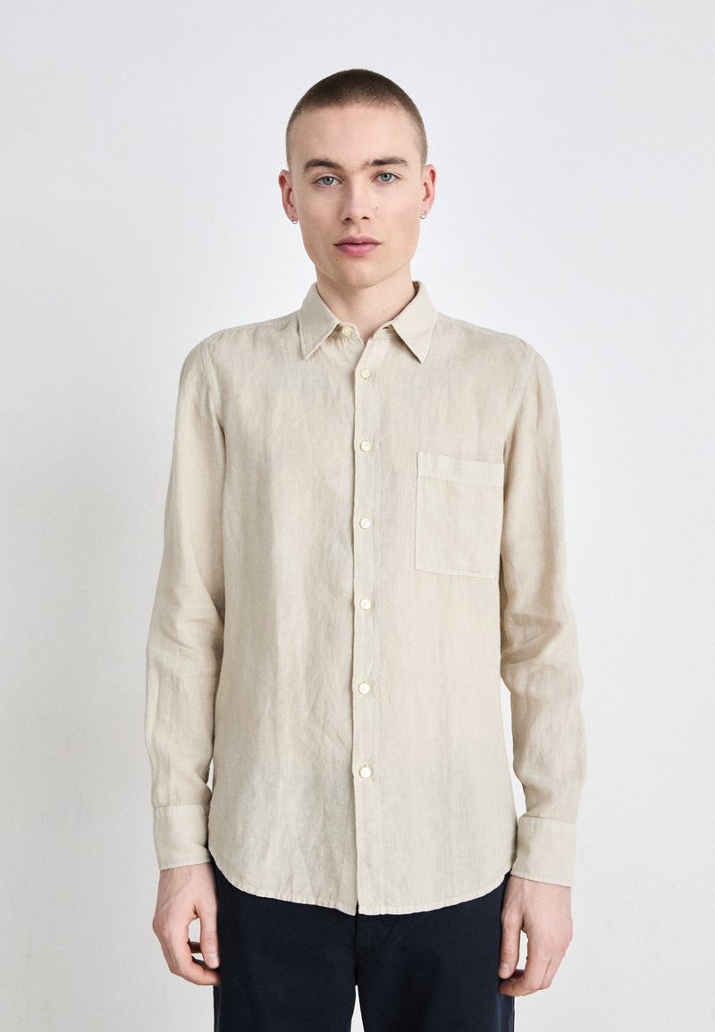 Camicia a maniche lunghe in lino beige con colletto, chiusura frontale a bottoni e taschino sul lato sinistro del petto. Presenta un orlo curvo e una texture minimale.