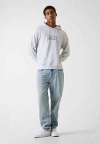 Sweat à capuche gris clair avec logo devant, capuche à cordon ; associé à un jean en denim bleu clair ; le mannequin est debout devant un fond blanc uni.