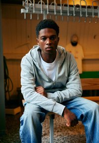 Grijze zip-up hoodie over een witte T-shirt, zittend op een houten bank, gecombineerd met vervaagde blauwe jeans. Korte krullende haren, serieuze uitstraling.