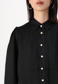 Black textured blouse featuring a collar, vertical pleats, and decorative pearl buttons down the front. Long sleeves with gathered shoulders.