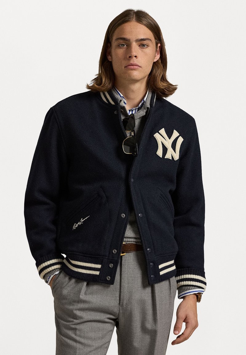 Marineblå uld varsity-jakke med cremefarvede striber, broderet NY-logo, ribbede manchetter og sidelommer, båret over en grå sweater og skjorte.