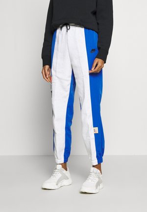 Persona con pantalones deportivos Nike azules y blancos con puños elásticos y zapatillas blancas, de pie frente a un fondo liso.