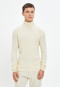 Mann in cremefarbenem, geripptem Rollkragenpullover und passenden Hosen steht vor einem weißen Hintergrund mit neutralem Ausdruck.