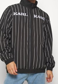 Svart randig jacka med hög krage, dragkedjestängning och ribbade ärmslut. Utmärkande vit text över bröstet. Lättviktsmaterial.