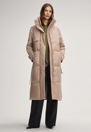 Donna con un lungo cappotto imbottito beige sopra una camicetta verde oliva e pantaloni neri a gamba larga, in piedi con le mani nelle tasche.