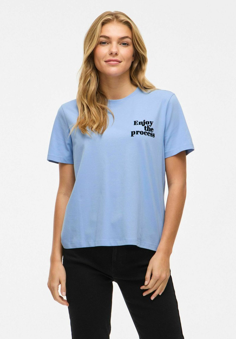 Ljusblå bomulls-t-shirt med korta ärmar, med svart text "Njut av processen" tryckt på vänster bröstparti. Platt fålldesign.