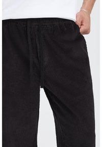 Pantalones jogger de pana negra con textura acanalada vertical, cintura elástica y cordón ajustable. Bolsillos laterales visibles.