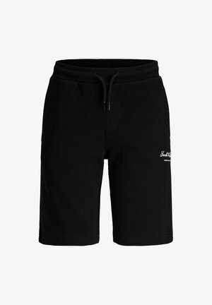 Niño sonriente de pie, vistiendo una camiseta gráfica azul marino, pantalones cortos negros, calcetines blancos y zapatillas deportivas blancas sobre un fondo claro y liso.