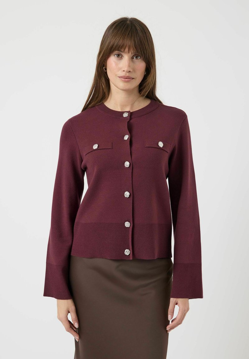 Cárdigan de punto burdeos con mangas largas, cuello redondo, dos bolsillos en el pecho y botones en tono plateado. Textura suave con una silueta ajustada.