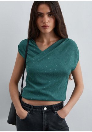 Mujer con cabello largo y oscuro lleva top sin mangas verde con textura y vaqueros negros, de pie con las manos en los bolsillos frente a un fondo liso.