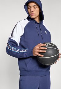 Μπλε φούτερ με κουκούλα σε ναυτικό μπλε χρώμα και λευκές λεπτομέρειες, με λογότυπο και διακοσμητική κορδέλα στα μανίκια. Μαλακή υφή με τσέπη καγκουρό.