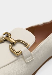 Λευκό μοκασίνι από δέρμα με υφή, με χρυσή μεταλλική λεπτομέρεια σε σχήμα σιδερόδρομου στη μύτη και ταμπά εσωτερική επένδυση.