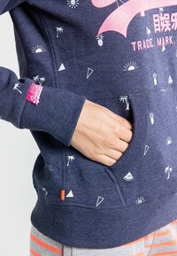 Felpa blu navy con motivi grafici bianchi, tasca a marsupio e patch logo rosa sulla manica. Tessuto morbido e dalla trama interessante. I disegni illustrativi includono palme e triangoli.