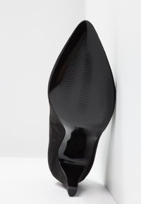Zapato de tacón alto negro con punta afilada, parte superior de ante y suela de goma lisa con estrías verticales. Etiqueta de tamaño visible.