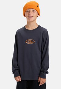 Langarmige marineblauwe shirt met oranje logo, gecombineerd met een oranje gebreide muts. Het shirt heeft een relaxte pasvorm en een zachte textuur.