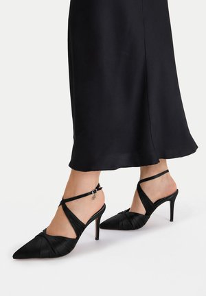 Frau trägt schwarze spitze High Heels mit Knöchelriemen und einem knielangen schwarzen Rock auf weißem Hintergrund.