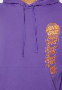Violetti huppari, jossa on kengurupocket ja oranssi graafinen teksti "Santa Cruz" pinottuna oikealla puolella.