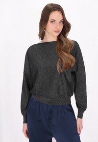 Schwarzer glitzernder Pullover mit weitem Ausschnitt, langen Ärmeln und breitem Bund, getragen über dunkelblauen Hosen, vor einem weißen Hintergrund.