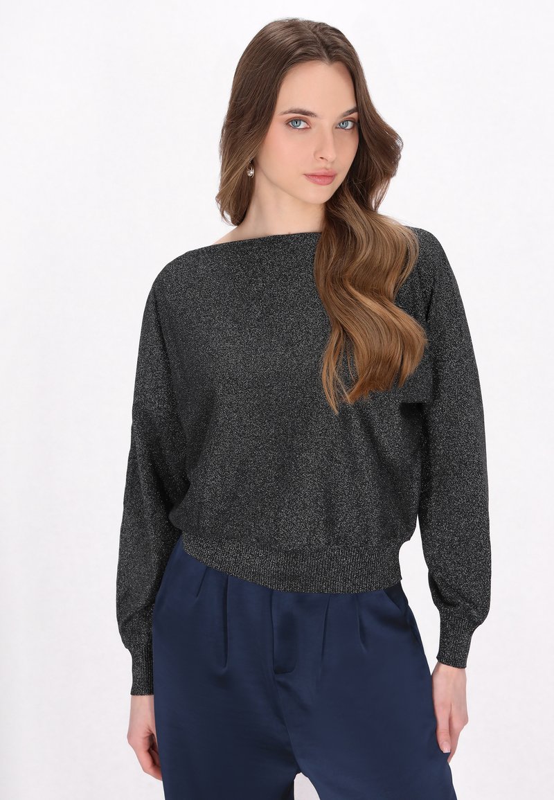 Schwarzer glitzernder Pullover mit weitem Ausschnitt, langen Ärmeln und breitem Bund, getragen über dunkelblauen Hosen, vor einem weißen Hintergrund.