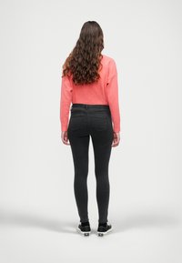 Jeans ajustados negros y un suéter coral con puños acanalados, combinado con zapatos negros. El cabello es largo y ondulado, visto desde atrás.
