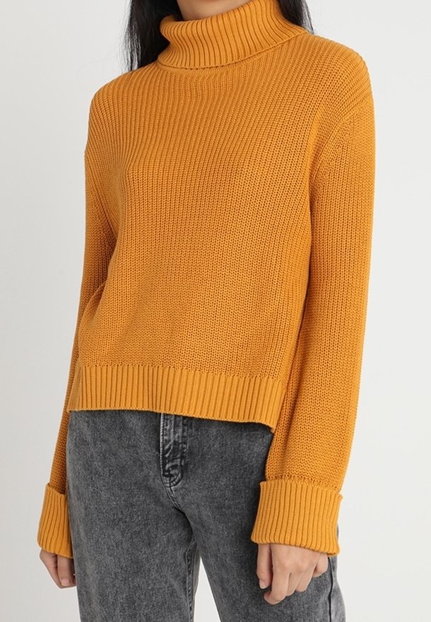 Femme portant un pull à col roulé moutarde côtelé avec manches retroussées et jean noir haute taille délavé.