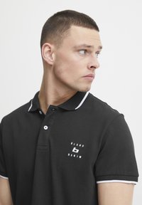 Černé polo tričko s bílým lemováním límce a dvouknoflíkovou légou. Obsahuje malé vyšívané logo na hrudi. Klasický střih a hladká textura.