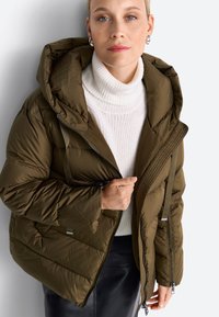 Femme portant une veste matelassée verte olive par-dessus un pull col roulé blanc et une jupe noire, tenant la fermeture éclair de la veste d'une main.