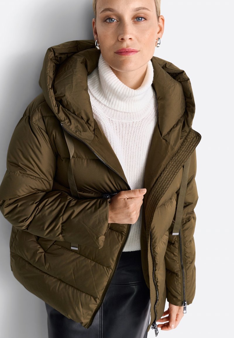 Femme portant une veste matelassée verte olive par-dessus un pull col roulé blanc et une jupe noire, tenant la fermeture éclair de la veste d'une main.