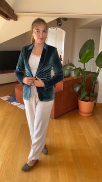 Tyrkysový sametový blazer s priliehavým dizajnom, biele tielko, svetlo šedé nohavice a šedé baletky. Vnútorné prostredie s kvetináčmi.