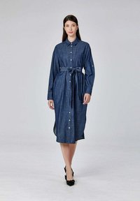 Femme portant une robe midi en denim boutonnée avec des manches longues et une ceinture à la taille, avançant sur un fond gris clair uni.