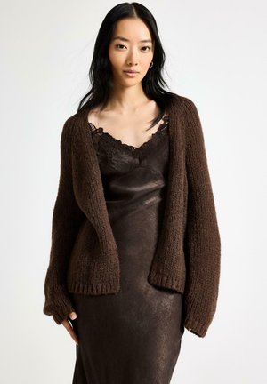 Kvinde iført en mørkebrun, blonde-trimmet kombinationskjole med en chunky strikket cardigan i matchende brun, stående mod en hvid baggrund.