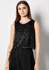 Top corto negro con lentejuelas y dobladillo ondulado, diseño sin mangas, superficie texturizada que refleja la luz, combinado con pantalones negros de tiro alto.