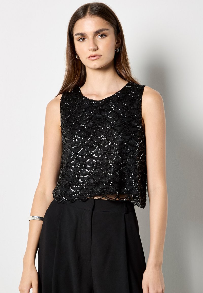 Top corto negro con lentejuelas y dobladillo ondulado, diseño sin mangas, superficie texturizada que refleja la luz, combinado con pantalones negros de tiro alto.