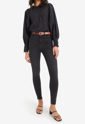 Zwarte blouse met geborduurd bloemenpatroon en ballonmouwen, gecombineerd met hoge donkere jeans en zwarte strappy hakken. Bruine riem als accent.