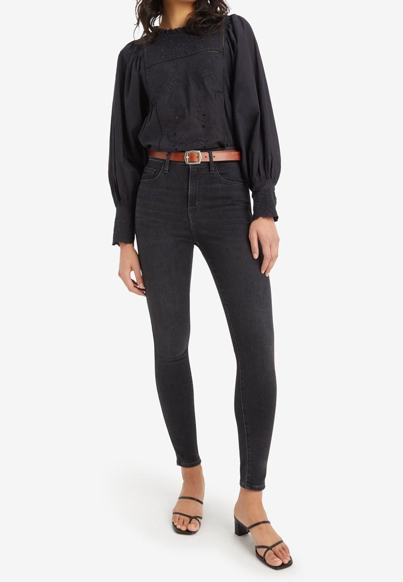 Blusa negra con diseño floral bordado y mangas globo, combinada con jeans oscuros de cintura alta y tacones negros con tiras. Cinturón marrón como acento.