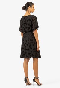 Robe noire à motifs floraux avec des manches transparantes, taille ajustée et jupe évasée. Se distingue par une texture lisse, un trou à clé au dos et une longueur au genou.