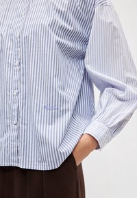 Personne portant une chemise ample rayée bleu et blanc à boutons, une main dans la poche d’un pantalon marron sur un fond uni.