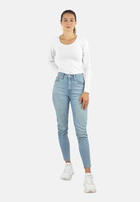 Weißes Langarmoberteil kombiniert mit hellblauen hoch sitzenden Jeans und weißen Turnschuhen. Das Outfit hat ein einfaches Design und eine schmale Passform.