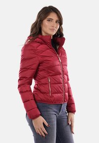 Giacca smanicata rossa con colletto alto, zip frontale e zip laterali. Presenta una texture trapuntata e un design aderente.