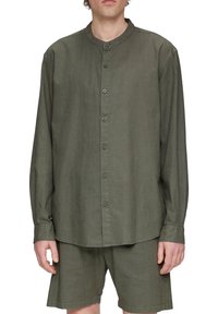 Man in een olijfgroen, met knopen gesloten, langemouw shirt met een bandkraag en bijpassende korte broek tegen een witte achtergrond.